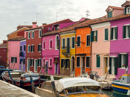 A row of brightly colored houses face the canal on the beautiful Venetian island of Burano, boats are docked on the canal in front of the houses - Burano, Venice, Italyの写真素材