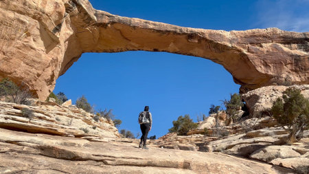 Female hiker walking up a rocky trail towards a natural sandstone formation known as Owachomo Bridge with a bright blue sky behind. Natural Bridges National Monument, Utah, USA.の写真素材