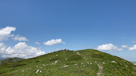A lush green mountain ridge under a bright blue sky with scattered clouds, featuring hikers enjoying nature. The serene landscape epitomizes outdoor adventures and tranquil natural beauty.の写真素材