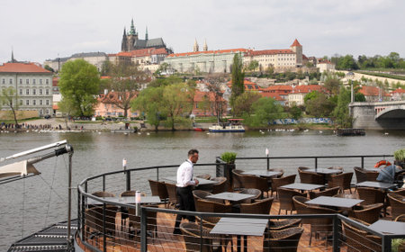 PRAGUE, CZECH REPUBLIC - APRIL 15, 2017: Waiter cleans tables at an outdoor restaurant on the riverbank of Vltava, with the Prague Castle in the backgroundのeditorial素材