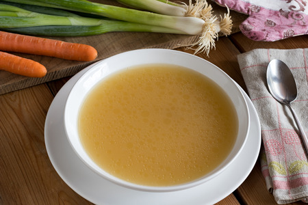 Bone broth made from chicken served in a white plate, with scallions and carrots in the backgroundの写真素材
