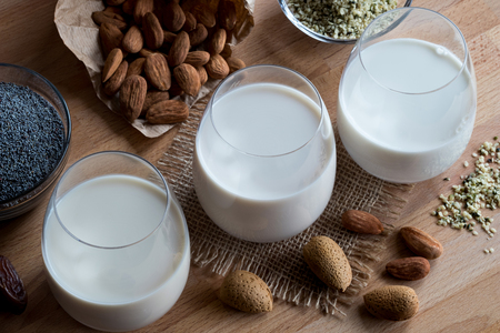 Three glasses of vegan plant milk - almond milk, poppy seed milk and hemp seed milk, with shelled and unshelled almonds, poppy seeds and hemp seeds on a wooden tableの写真素材