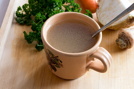 Chicken stock in a vintage mug with a spoon, with parsley, carrot and celery root in the backgroundの写真素材