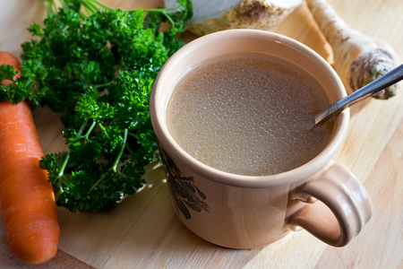 Chicken stock in a mug on a wooden table, with parsley, carrot and celery root in the backgroundの写真素材