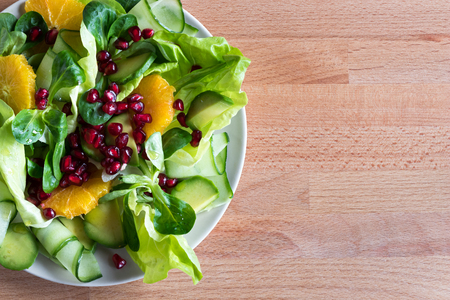 Vegetable salad (lettuce, corn salad, cucumber, avocado, orange, pomegranate) on a wooden background with copy space, top viewの写真素材