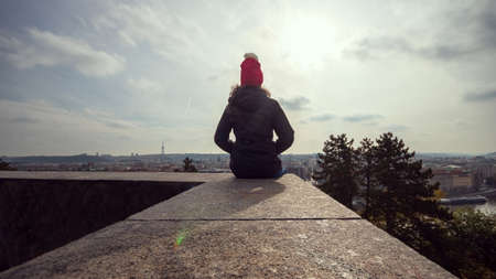 Woman in winter jacket sitting on a wall in the Letna park and looking at the panorama of Prague