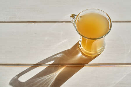 Bone broth in a glass cup on a white table, with a shadow from the sun