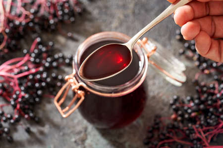 Hand holding a spoon with homemade black elder syrup, with fresh elderberries in the backgroundの写真素材