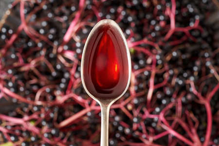 Black elder syrup on a spoon with fresh elderberries in the background, top view