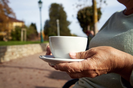 Hand of an elderly woman holding a white cup and drinking coffee outdoors on a sunny autumn dayの写真素材