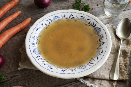 Chicken bone broth or soup in a white plate with blue ornaments on a table, with fresh vegetablesの写真素材