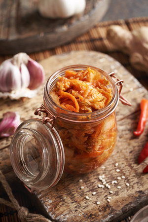 Fermented kimchee in a glass jar on a table with chilli peppers, sesame seeds, ginger and garlic on a tableの写真素材