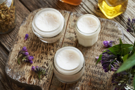 Three jars of homemade comfrey root ointment made with rendered lard with fresh blooming Symphytum plant on a tableの写真素材