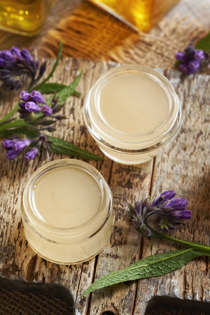 Two jars of homemade comfrey root ointment made with hot rendered pork lard and fresh symphytum plant on a tableの写真素材
