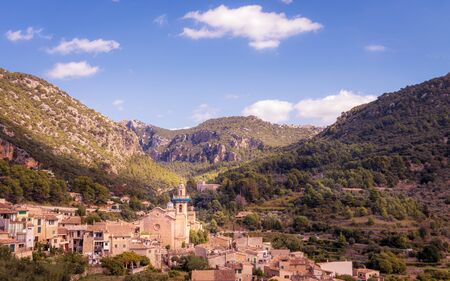 Valldemossa church and surrounding town with mountains, forests and blue sky with white clouds,...