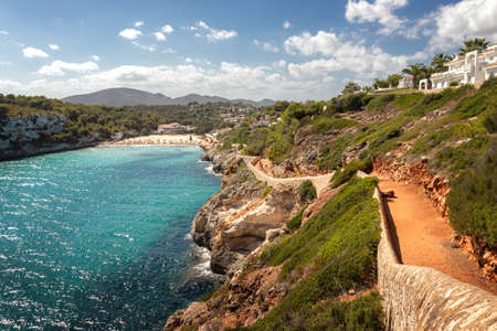 Typical Spanish beach scene with pathway leading to beach surrounded by green hills and mountains with blue sky, Cala Romantica, Mallorcal, Spain.の写真素材