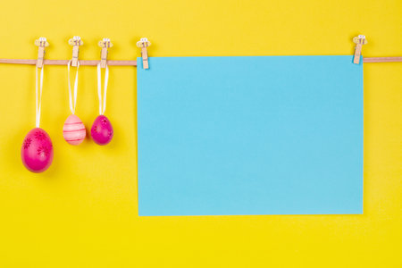 Blank blue sheet of paper and three pink Easter eggs hang at rope on yellow background. Easter greeting card concept with designated writing spaceの写真素材