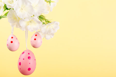 Delightful depiction of Easter joy featuring three pink eggs gracefully suspended from a blooming branch against yellow pastel backgroundの写真素材