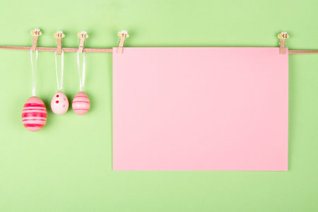 Blank pink pastel sheet of paper and three pink Easter eggs hang at rope on light green pastel background. Easter greeting card idea with blank space for your wordsの写真素材