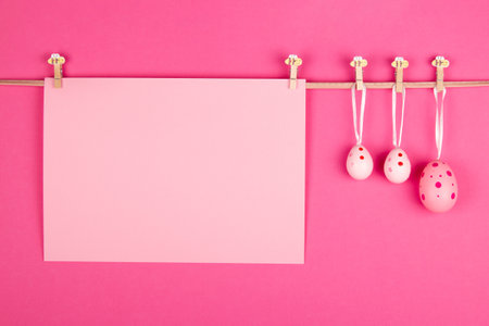 Blank pastel pink sheet of paper and three pink Easter eggs hang at rope on vibrant pink background. Easter card design with open area for personalized messageの写真素材