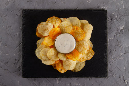 Top view of heap of potato chips with creamy white dipping sauce on black slate serving board on gray plaster table. Deliciously Savory Chips and Dipの写真素材