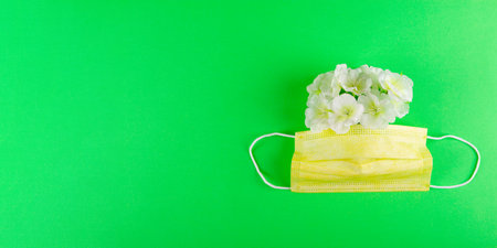 Yellow medical mask and white flowers on vibrant green background with copy space at the left side....