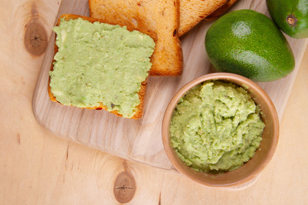On pale yellow wooden surface you'll find still life with avocado toast, luscious avocado spread and two perfectly fresh avocado fruits close-upの写真素材