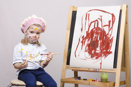 Portrait of a three-year old girl playing in the artist. Girl mud paints. Standing behind you easelの写真素材