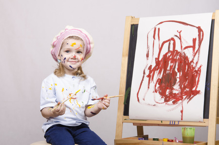 Portrait of a three-year old girl playing in the artist. Girl mud paints. Standing behind you easelの写真素材