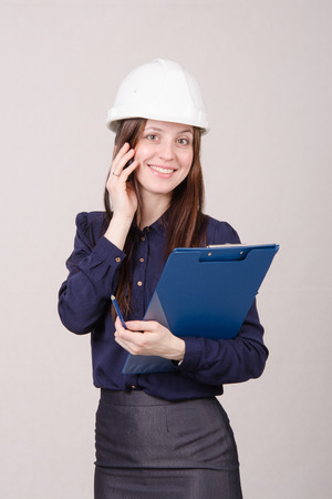Young woman builder in helmet standing with a folder in hand and talking on phoneの写真素材