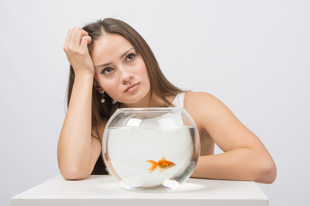 A young girl sits next to a round aquarium in which swimming goldfishの写真素材