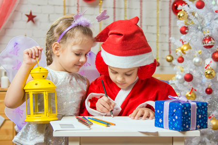 A girl dressed in a red suit Santa Claus draws pencils sitting at the table, next is a girl dressed as a fairy with the candlestickの写真素材