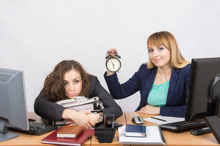 Two girls in the office at the end of the day, one with a smile, holding a clock, another weary lies on foldersの写真素材