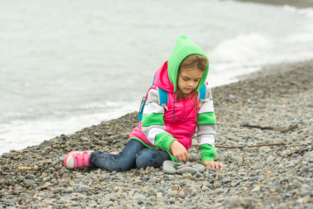 Seven-year girl sitting on a pebble beach in the warm clothing and with enthusiasm plays with stonesの写真素材
