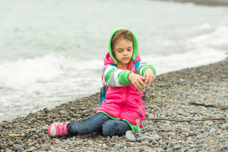 Seven-year girl sitting on a pebble beach in the warm clothing and pours out through her fingers small stonesの写真素材