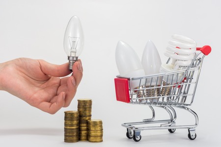 In the grocery cart are LED and energy-saving light bulbs, next to a hand is holding an incandescent lamp, there is a stack of coinsの写真素材