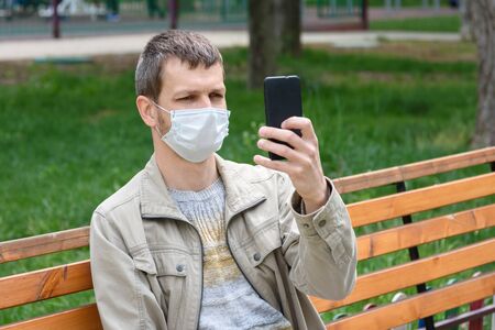 A man in a medical mask sits on a bench and photographs himselfの写真素材
