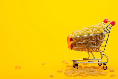 A grocery cart to the top is filled with pasta on a bright orange background, some pasta is lying next to the cartの写真素材
