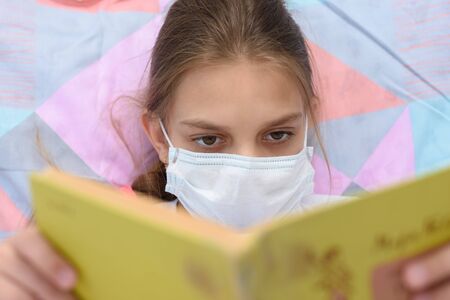 Girl in a medical mask reads a book while lying in bed, close-upの写真素材