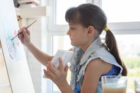 Girl diligently draws a picture on an easel in front of a large window