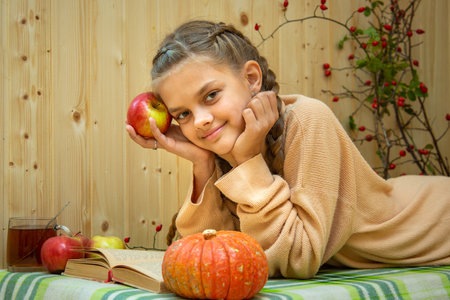 A girl lying down reads a book, a pumpkin and apples lie nearby, a girl joyfully looks into the frameの写真素材