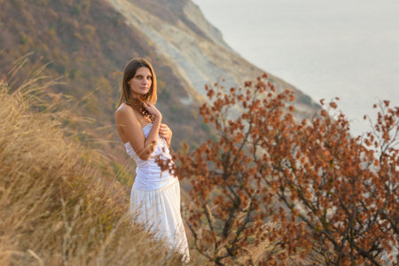 Beautiful girl in white dress hugging wild flowers on the background of beautiful sceneryの写真素材