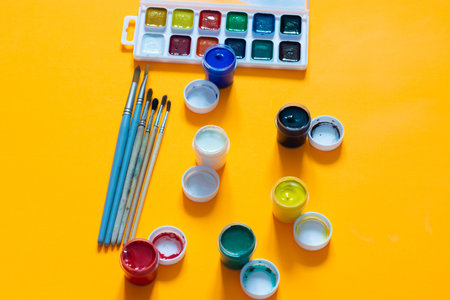 Brushes, jars of gouache and watercolor paints lie on a yellow backgroundの写真素材