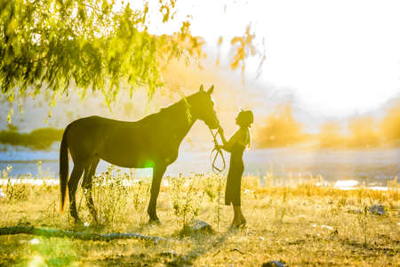 A girl stands in front of a horse on the shore of a lake and is flooded with warm sunbeams at sunsetの写真素材