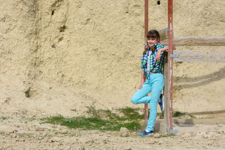 Stylish cute girl in a plaid shirt and jeans leans on a wooden structure against a background of a rockの写真素材