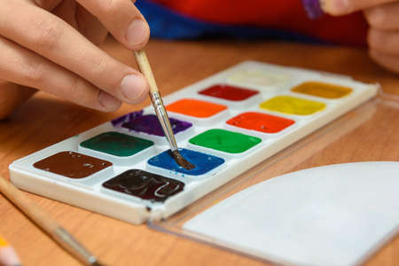 The child's hands are dipping with a brush in the desired color of watercolor paintsの写真素材
