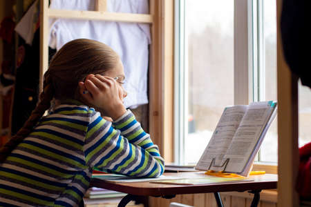 The schoolgirl carefully reads the task in the textbook while sitting at the table by the window at homeの写真素材