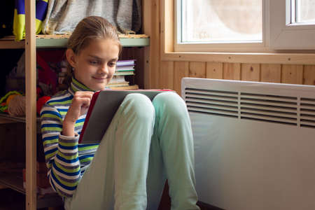 A girl plays in a tablet sitting next to a radiator by a window in a country houseの写真素材
