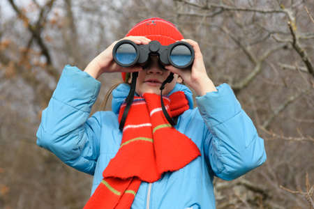 A girl looks through binoculars on a background of a winter forest, front view, close-up