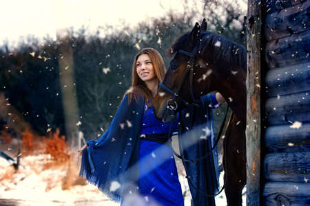 Beautiful girl in a blue stole stands next to a horse near wooden buildings on a snowy winter dayの写真素材